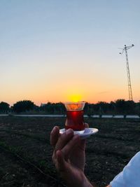 Cropped hand holding turkish tea on field against sky during sunset