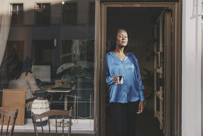 Female business professional holding coffee cup while standing with eyes closed at office doorway