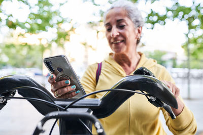 Elderly female athlete using smartphone to rent electric bicycle after training in sunny morning on street