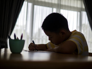 Surface level of boy drawing on table while sitting against window at home