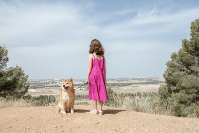 Back view of unrecognizable little girl in dress standing with cute obedient purebred shiba inu dog on hill while spending summer day together in countryside
