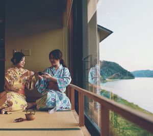 Women sitting on railing by sea against mountain