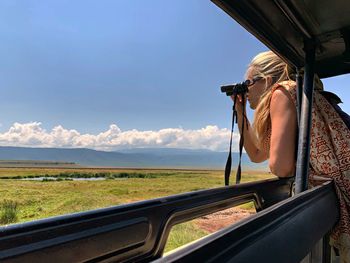 Side view of woman looking through binoculars while standing in jeep against sky