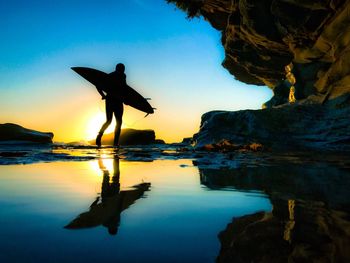 Rear view of silhouette man holding surfboard while walking on shore against sky during sunset