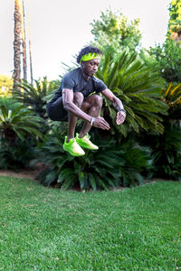 Full body of young african american male athlete jumping high above green lawn while working out in tropical park