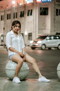 Full length portrait of young woman sitting by street on bollard in city at night