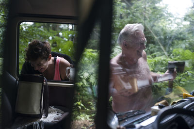 Mid adult man reflecting on mirror while senior person seen through windshield in forest