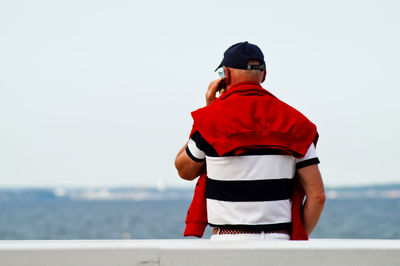 Rear view of man talking on phone while standing at railing against sea