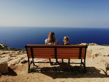 Rear view of siblings sitting on bench at cliff by sea