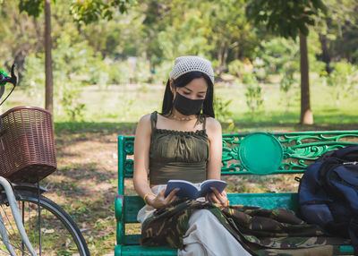  beautiful asian woman  with face mask sitting and reading a book on a wooden bench in a public park