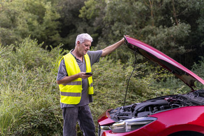 Senior man standing at his broken car wearing a safety vest and using his smartphone