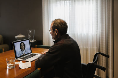 Disabled senior man sitting on wheelchair while female doctor consulting through video call on laptop