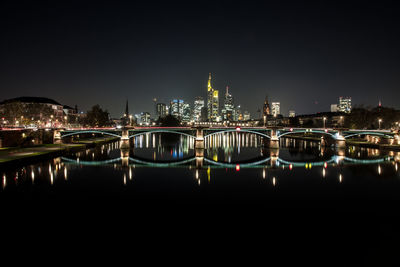 Illuminated bridge over river by buildings against sky at night