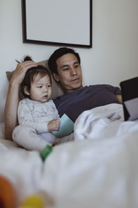 Father using digital tablet by male toddler in bedroom