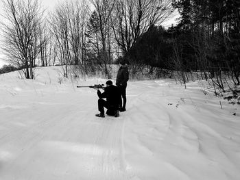 Man shooting with rifle by friend on snow covered field against bare trees