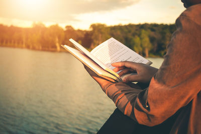 Midsection of man holding book against sea during sunset