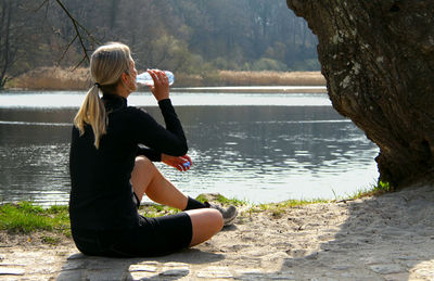 Rear view of mid adult woman drinking water while sitting at lakeshore in forest