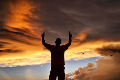 Rear view of man with arms raised standing against cloudy sky during sunset