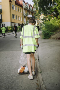 Full length rear view of teenage boy wearing reflective clothing while walking with garbage bag on street