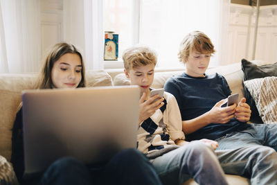 Girl using laptop and boys text messaging on mobile phone while sitting at sofa