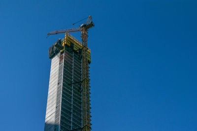 Low angle view of crane by building against clear blue sky