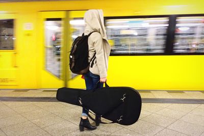 Side view of woman in hooded jacket holding guitar while waiting at railway station