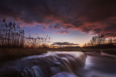 Flowing river stream in field against sky during sunset