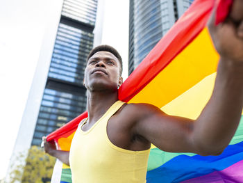 Thoughtful young man holding rainbow flag in front of modern buildings