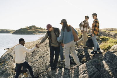 Young man helping male friend in climbing hill while hanging out with each other at sunny day