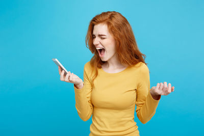 Angry young woman screaming with smart phone against blue background