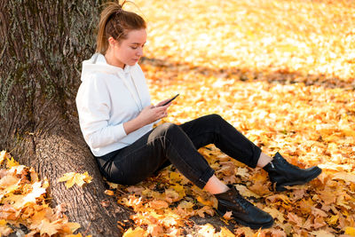Young woman using mobile phone while sitting on autumn