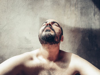 Portrait of shirtless young man with eyes closed against wall