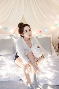 Portrait of young woman sitting with toy by illuminated lighting equipment on bed