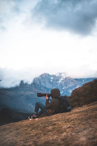 Side view of traveling photographer with backpack taking photo sitting on ground in mountains