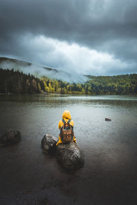 Rear view of person sitting on rock in lake against mountain and sky