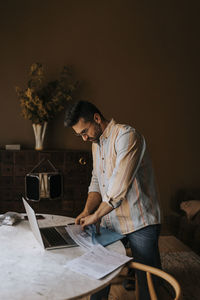 Man sorting out documents near laptop on table while standing at home