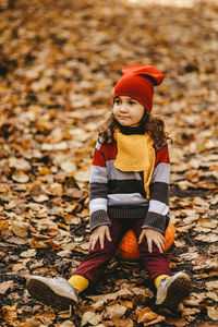 Happy cute little child in warm bright clothes sitting on a pumpkin in the autumn forest outdoors