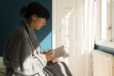 Woman wearing bathrobe holding cup while reading book in bedroom at home