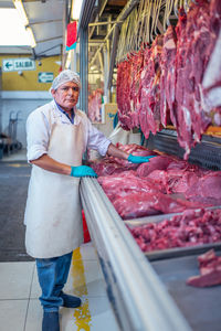 Mature man in uniform touching fresh meat on stall while working in butchery on market