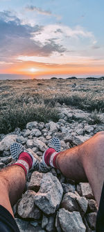 Low section of man relaxing on beach against sky during sunset