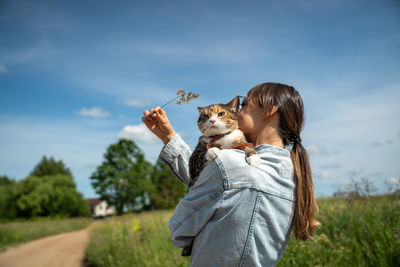 Rear view, woman holding serious cat in arms, trying to play, interest pet in twig of plant.