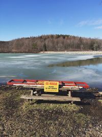 Information sign with ladder on bench at lakeshore against sky