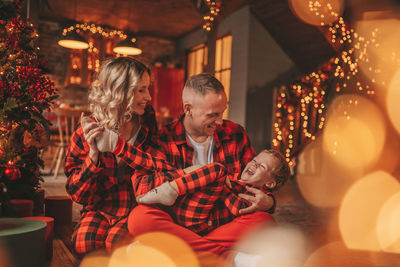 Smile active parents with small son in red checkered sleepwears waiting santa indoor.