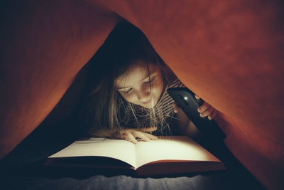 Close-up of girl with mobile phone light reading book under blanket