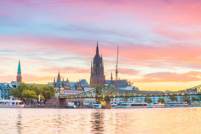 Bridge over river by buildings against sky during sunset