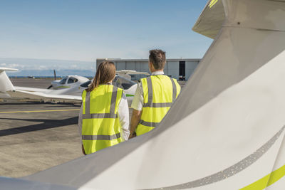 Rear view of engineers wearing reflective clothing standing against blue sky at airport runway