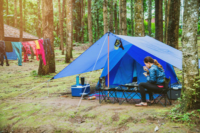 Woman eating food while sitting on chair by tent in forest