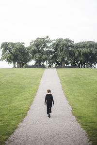Rear view of young woman walking amidst grassy field against trees and sky