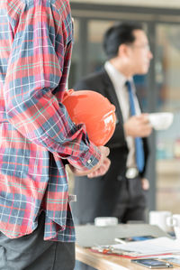 Midsection of architect standing holding hardhat with businessman standing in office