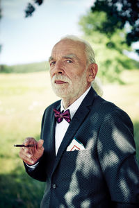 Portrait of senior man wearing suit coat, bow tie and dress handkerchief in nature smoking cigarillo